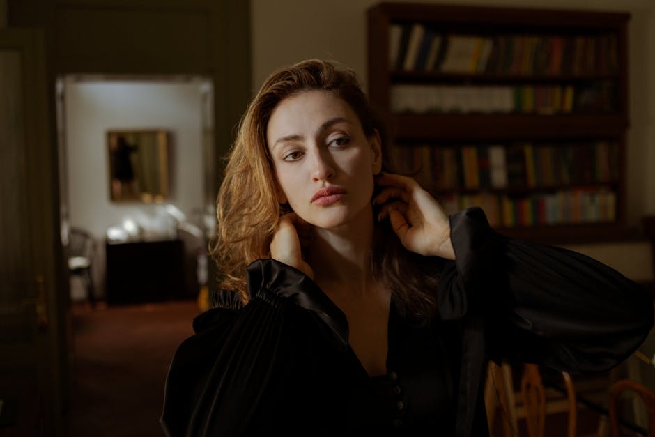 A woman with wavy hair wearing a dark outfit poses thoughtfully indoors, standing near a bookshelf and a doorway.