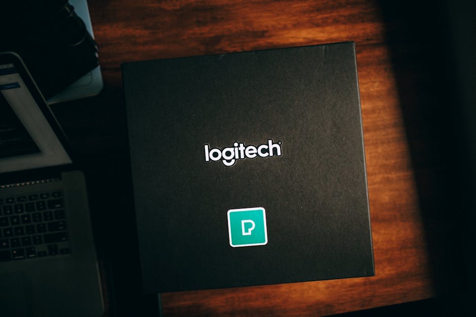 The image depicts a large black box with the 'Logitech' logo prominently displayed on the top. The box is placed on a wooden surface beside a laptop, hinting at a possible tech or electronics theme. The presence of the logo suggests it could be packaging for a Logitech product. The lighting is warm, creating a cozy ambiance.
