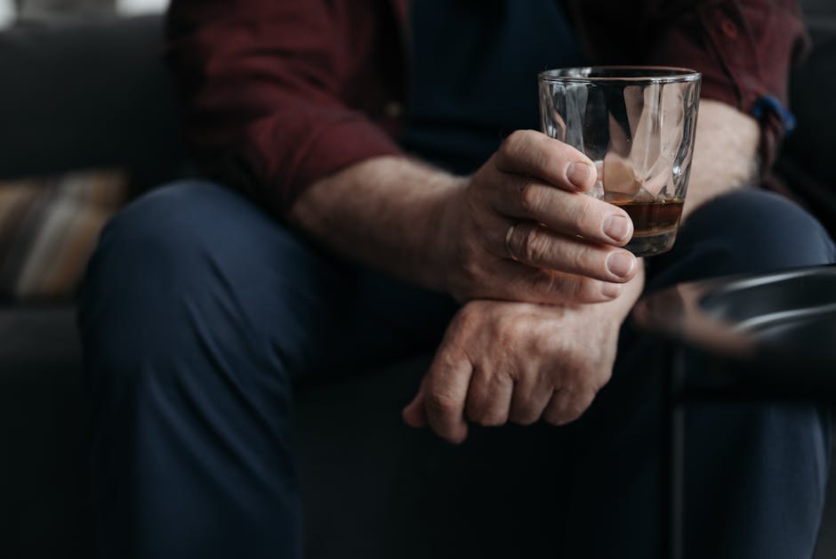 A close-up image of a man seated on a couch, holding a glass of whiskey in one hand. The focus is on the man's hand and the drink, with a slightly blurred background that includes parts of his clothing and the couch. The atmosphere is calm and relaxed.