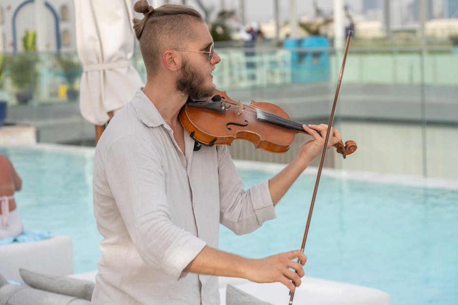 A man with a light beard and sunglasses is playing a violin outdoors near a swimming pool. He is wearing a light-colored, long-sleeved shirt, and has his hair tied up. The background includes poolside furniture and an outdoor setting, suggesting a relaxed atmosphere with focus on the musical performance.