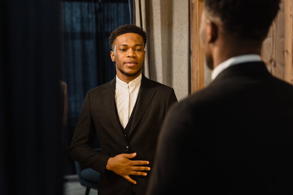 A man wearing a formal black suit and white shirt stands in front of a mirror, examining his appearance. The reflection shows the back of his head, highlighting the suit's fit and style.