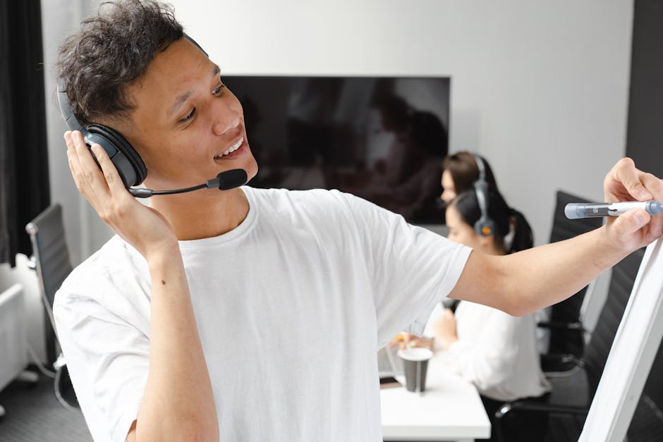 A man wearing a white T-shirt and a headset is standing in front of a whiteboard, writing or drawing with a marker. He appears to be teaching or conducting a training session. In the background, there are two people, also wearing headsets, sitting at a table with a laptop. The setting is likely an office or a training room.