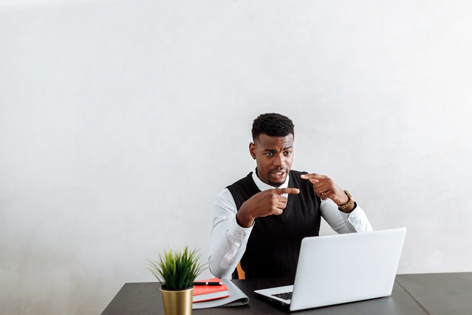 A man sitting at a desk engaged in a video call on his laptop. He is gesturing with his hands, indicating an active conversation. The desk has a small plant and a few notebooks. The background is plain, emphasizing the focus on the man and his interaction through the laptop.