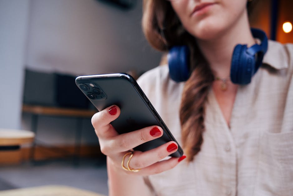 A person wearing blue headphones holds a smartphone with red-painted nails visible.