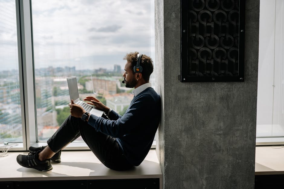 A man with headphones sits next to a large window working on a laptop in a modern office space.