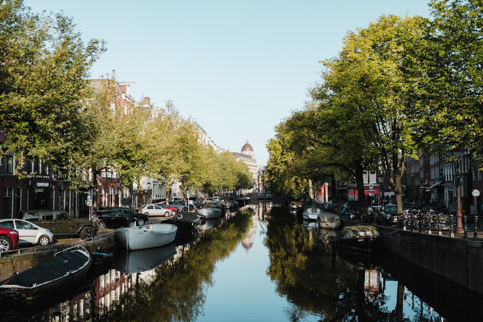 A serene canal lined with boats and bordered by trees and buildings, with a distant dome visible in the background.