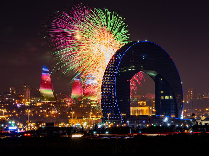 A vibrant nighttime cityscape featuring large, colorful fireworks exploding behind a distinctive arch-shaped building.