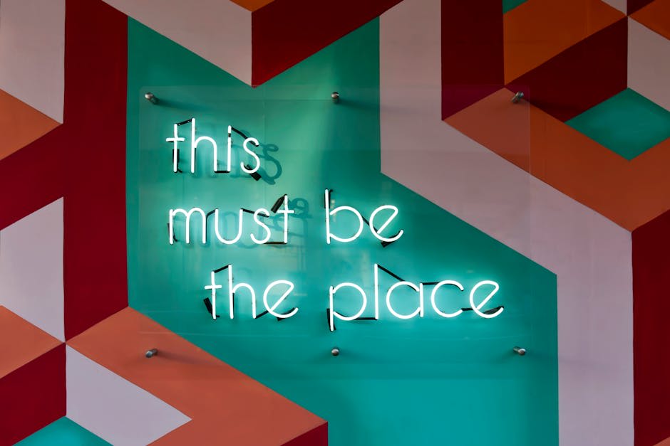 A neon sign displaying the words 'this must be the place' mounted on a colorful geometric patterned wall. The background features teal, orange, and red shapes, providing a vibrant and artistic atmosphere.