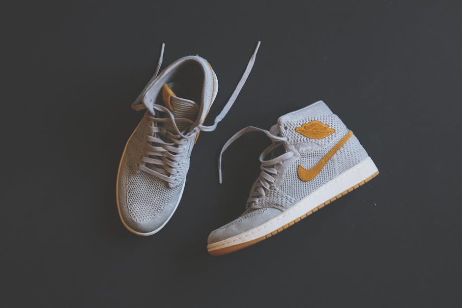A pair of grey Nike high-top sneakers with orange details and white soles, displayed on a dark background. The sneakers are positioned with one lying flat and the other standing upright. The image highlights the design and color contrast of the shoes.