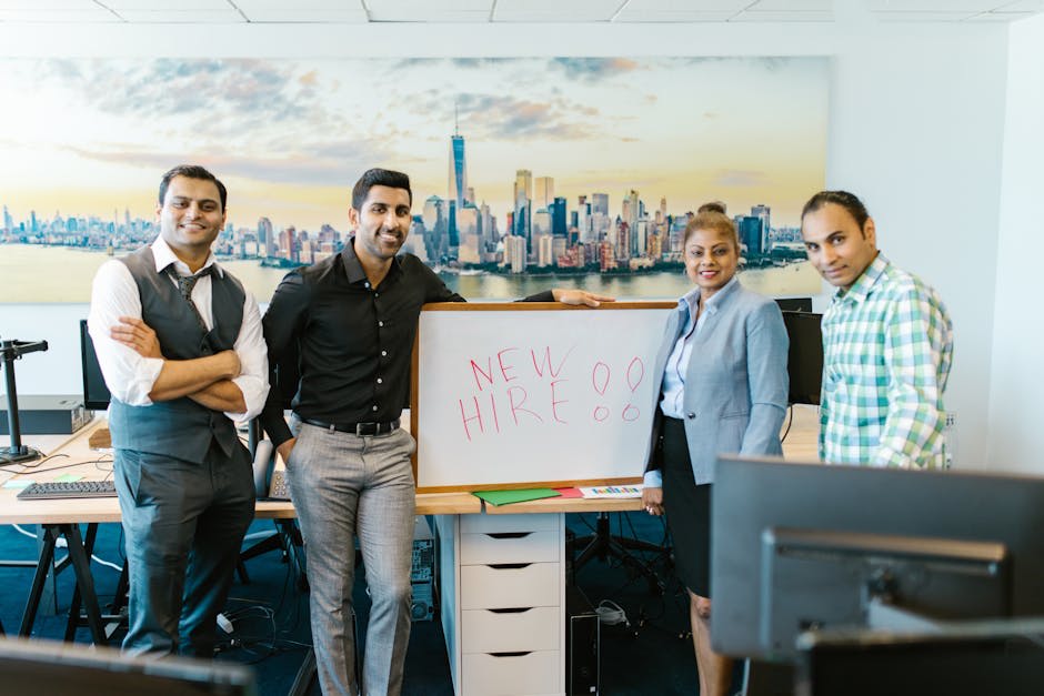 A group of four people standing in an office environment, smiling and posing around a whiteboard with the words 'New Hire' written on it. The background shows a large cityscape image on the wall. The image conveys a welcoming atmosphere for a new employee in a professional setting.