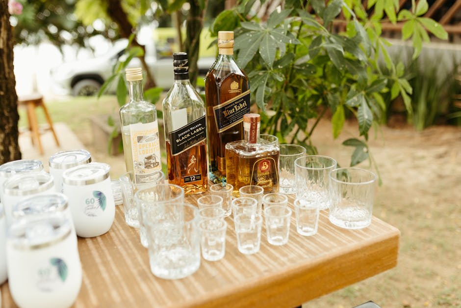 This image shows a variety of alcoholic beverage bottles, including whiskey and rum, displayed on a wooden table outdoors. The setup includes various sizes of empty glasses arranged next to the bottles. The scene suggests a casual outdoor gathering or party setting, surrounded by greenery.