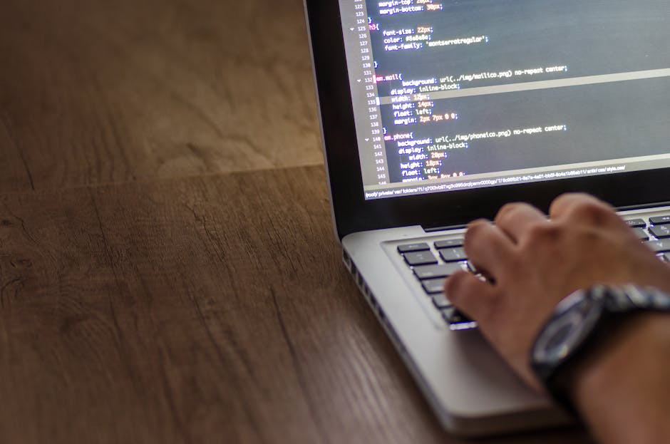 A person is typing on a laptop displaying lines of code. The laptop is on a wooden surface. The focus is on the coding process, and the screen shows HTML and CSS code.