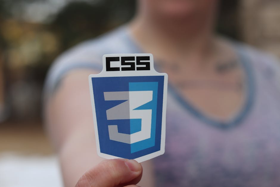 A person holding a CSS3 logo sticker prominently in the foreground. The sticker features the CSS3 emblem in blue and white. The focus is on the sticker, with the person intentionally blurred in the background.