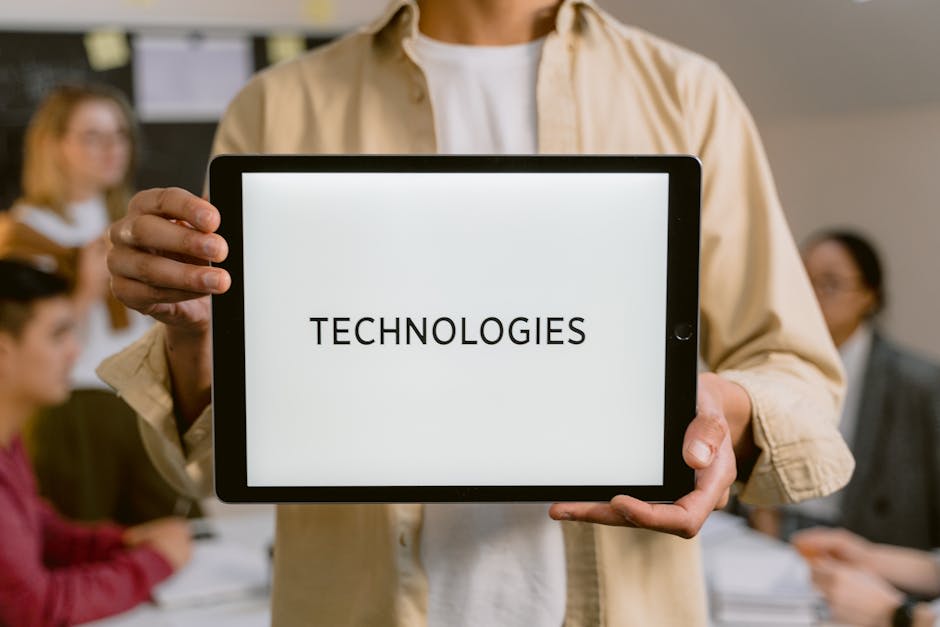 A person is holding a tablet that displays the word 'TECHNOLOGIES' in a bold font. The background features a group of people in an office setting, indicating a focus on technology in a business or collaborative environment. The image highlights modern technological themes and teamwork.