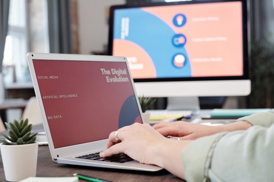 A person using a laptop with a presentation on 'The Digital Evolution' displayed on the screen. The laptop screen shows topics like social media, artificial intelligence, and big data. A desktop monitor in the background also displays related digital content. The setting suggests a work or study environment focused on technology and digital transformation.