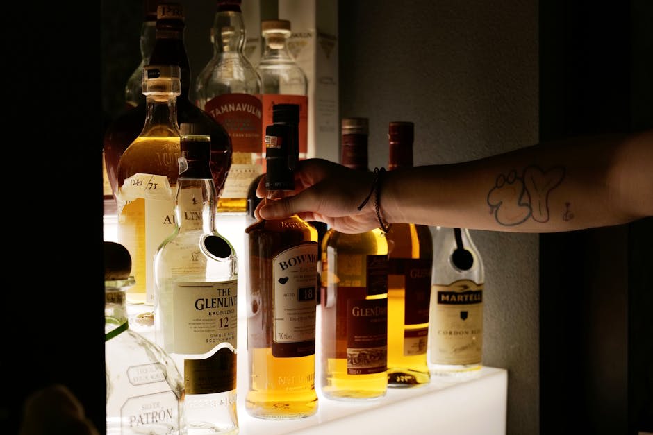 A person selecting a whisky bottle from a shelf displaying various liquor bottles. The arm has a tattoo, and the shelf includes brands like Glenlivet and Bowmore, set against a dimly lit background.
