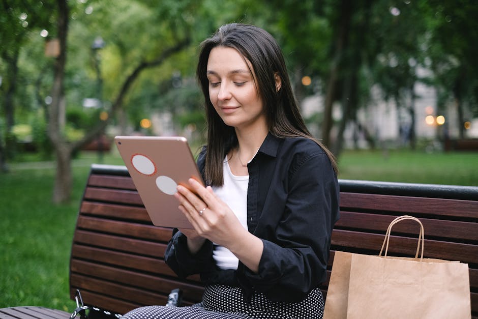A woman sitting on a park bench looks at a tablet while a paper shopping bag is placed next to her.