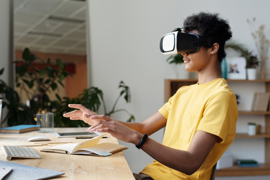 A person wearing a virtual reality headset, sitting at a desk with a keyboard, notebook, and glass of water. The person appears to be interacting with a virtual environment, indicated by their hand movements. The setting includes indoor plants and shelves in the background, suggesting a home or office environment. The person is wearing a yellow shirt.