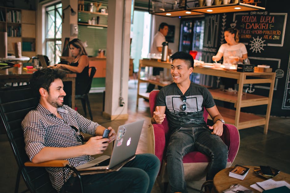 Two men are sitting and conversing in a relaxed café setting, one using a laptop and the other holding a smartphone, while other people work in the background.