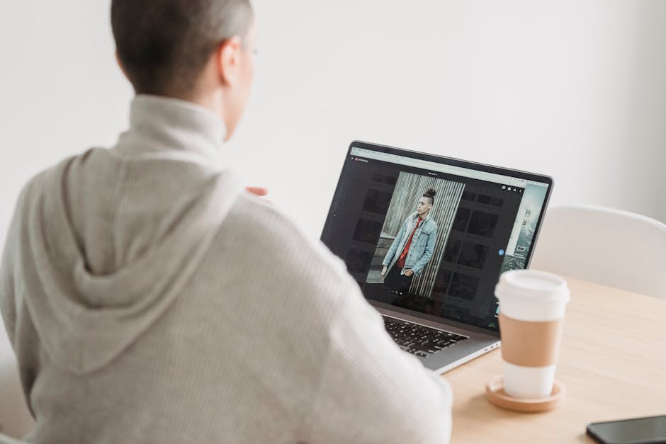 A person in a gray hoodie sits at a wooden table, working on a laptop. The laptop screen displays an image of a casually dressed person standing against a striped wall. A take-out coffee cup is placed on the table next to the laptop. The setting appears to be a bright and minimal workspace.