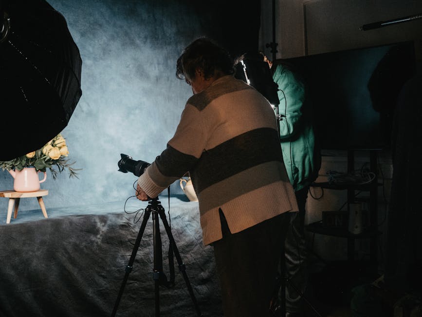 A person is setting up a camera on a tripod in a studio, focusing on a floral arrangement under studio lighting.
