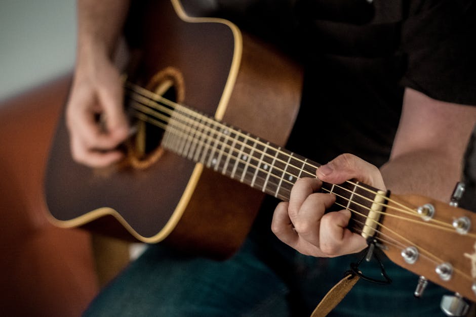 A close-up image of a person playing an acoustic guitar, focusing on the hands strumming and pressing the strings. The background is blurred, emphasizing the details of the guitar and the musician's hands.