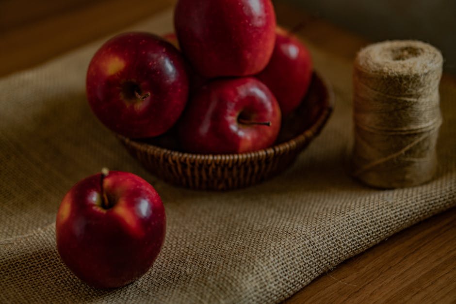 A rustic still life image featuring a woven basket filled with several red apples placed on a piece of jute fabric. One apple is positioned outside the basket on the jute, and there is a coil of thick twine next to the basket. The setting has a cozy and natural aesthetic, suitable for themes related to fruit, rustic decor, or autumn.