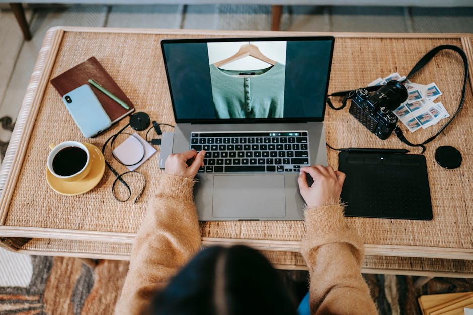 A person is working on a laptop displaying an image of a shirt, surrounded by items like a camera, coffee, a notebook, and a smartphone on a wicker desk.