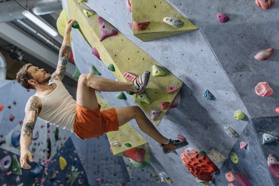 A man in a tank top and shorts is rock climbing indoors on a bouldering wall with colorful grips.