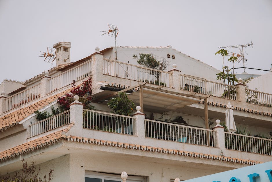 A beige, multi-level house with a terracotta-tiled roof, balcony railings, plants, and several rooftop antennas.