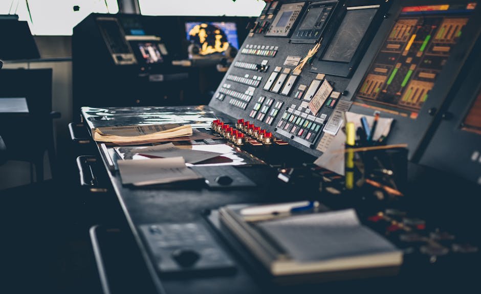 Image of a ship's control room with a variety of electronic devices, control panels, and papers scattered on the desk. The control room features multiple screens and buttons, indicating a complex and technical workspace.