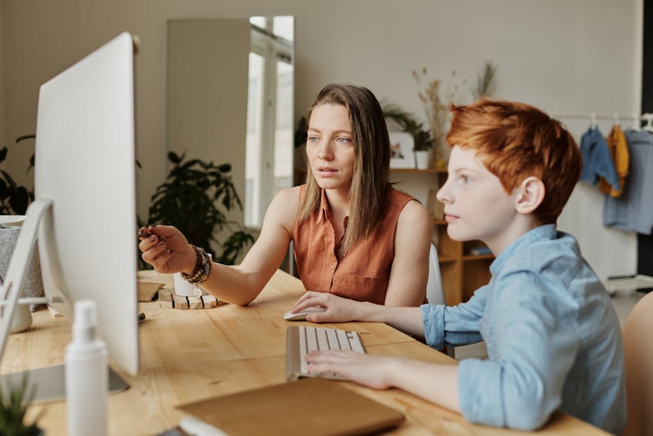 A woman and a child are sitting at a desk in front of a computer, with the woman pointing at the screen, seemingly explaining something.