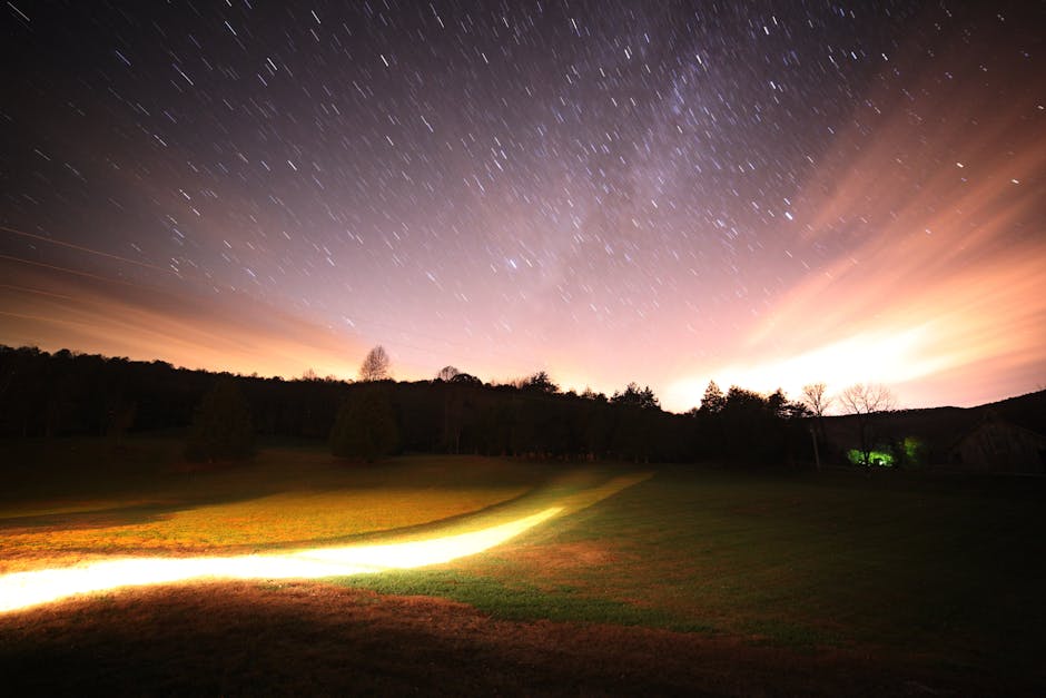 A long-exposure nighttime photograph of a grassy field with a light trail in the foreground and a starry sky above a distant tree line.