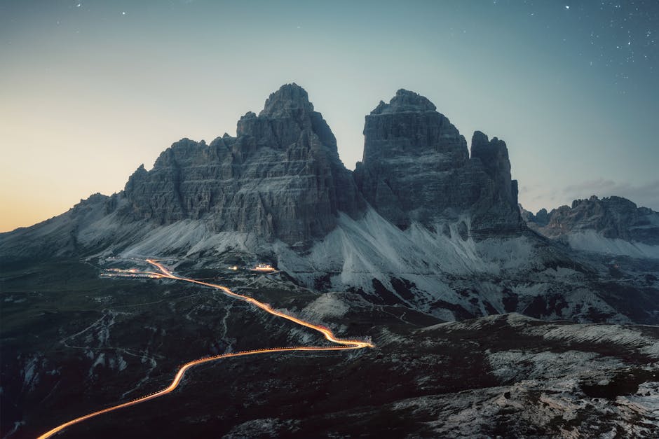 A dramatic view of the Tre Cime di Lavaredo mountain peaks at dusk, with a winding road illuminated by streaks of light in the foreground.