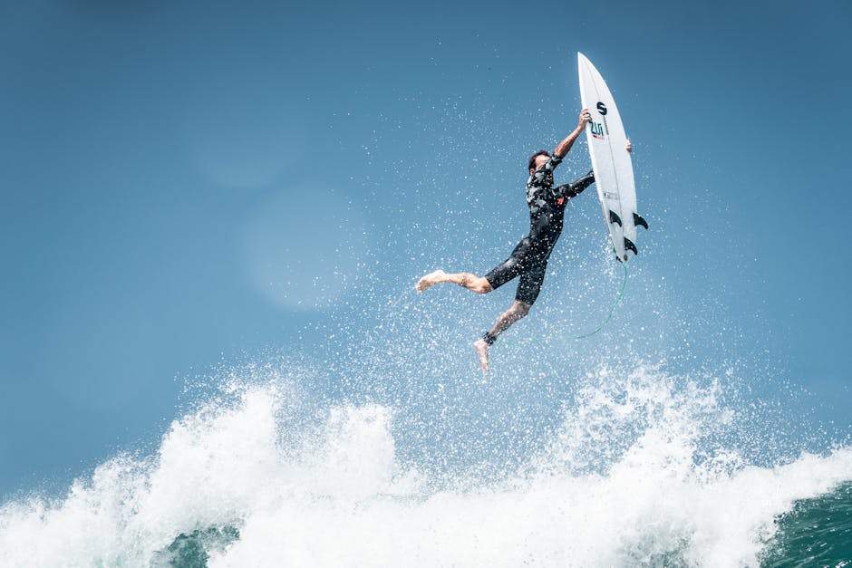 A surfer in a wetsuit is airborne above a wave while holding their surfboard against a clear blue sky.
