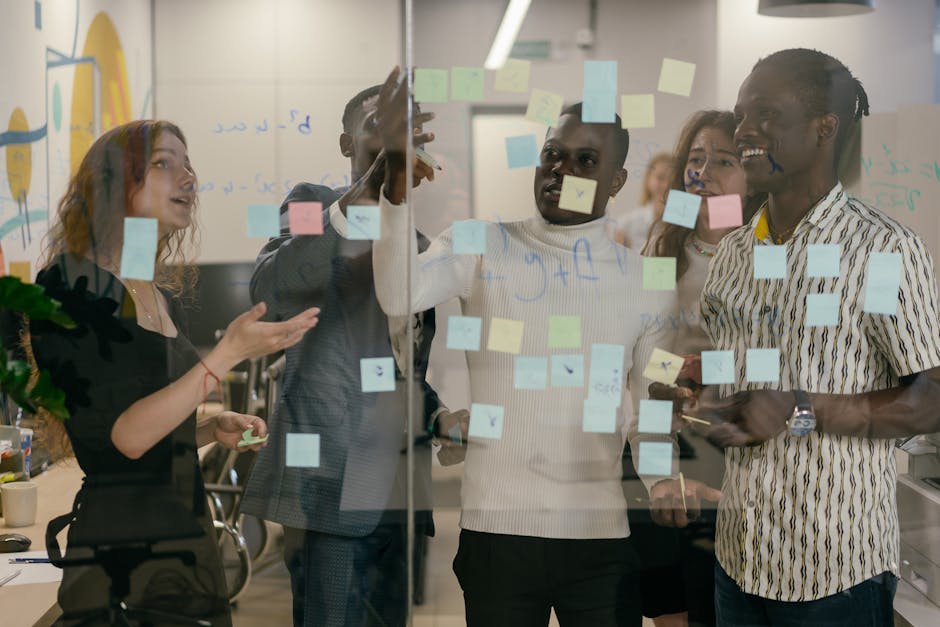 A group of people collaborate by discussing and organizing sticky notes on a glass board in an office setting.
