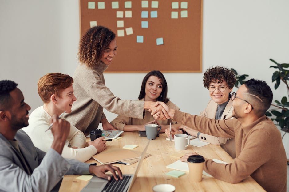 A diverse group of colleagues having a meeting at a conference table. A woman with curly hair is shaking hands with a man with glasses across the table. Other team members are seated around, smiling, with notepads and coffee cups on the table. Background features a bulletin board with post-it notes and plants.