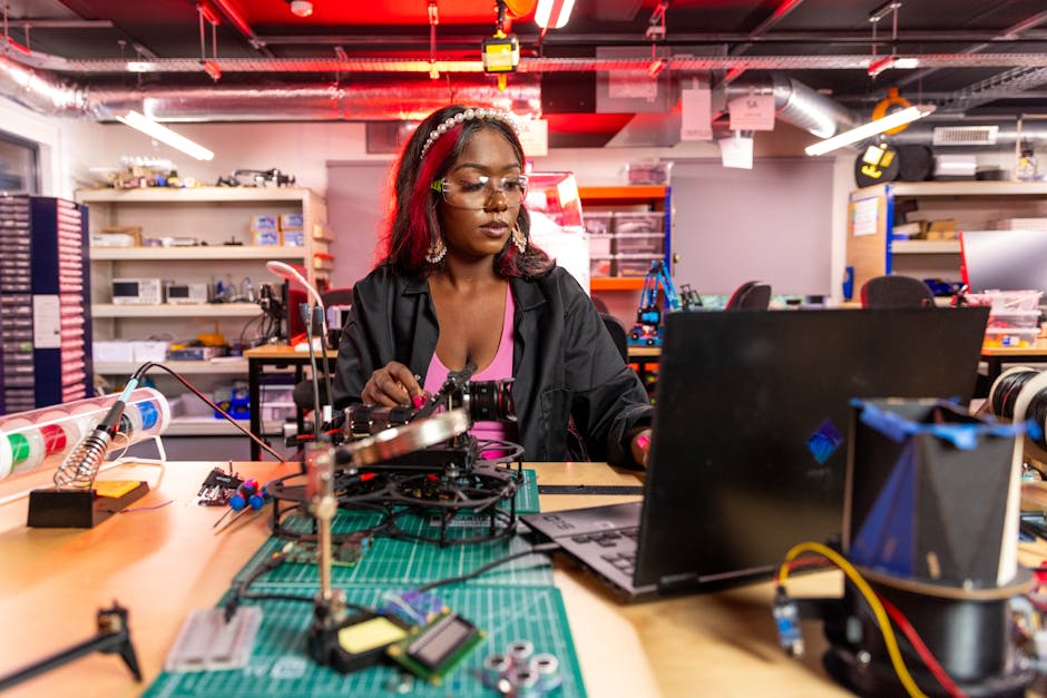 A woman wearing safety glasses works on an engineering project at a desk with electronic components and a laptop in a lab setting.