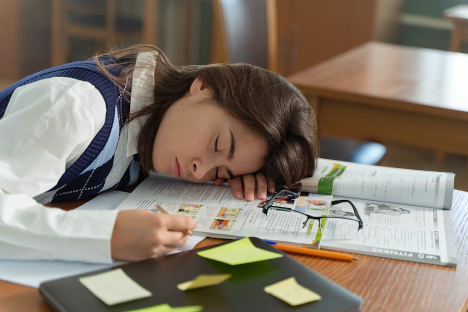 A person has fallen asleep on an open textbook at a desk, surrounded by study materials including a notebook, glasses, and sticky notes.