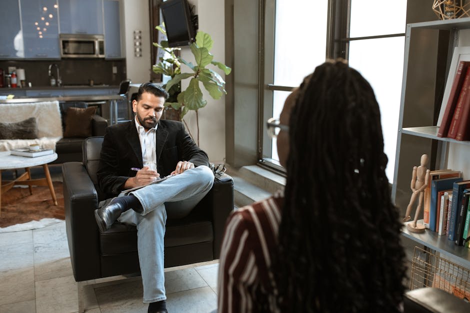The image shows a therapy session in progress. A man, possibly a therapist, is sitting in an armchair, taking notes, while a woman faces him, partially out of frame, appearing to be speaking or listening. The setting resembles a comfortable room with a kitchen in the background, a plant, and bookshelves filled with books, suggesting a private office or a cozy space designed for counseling or therapy conversations.