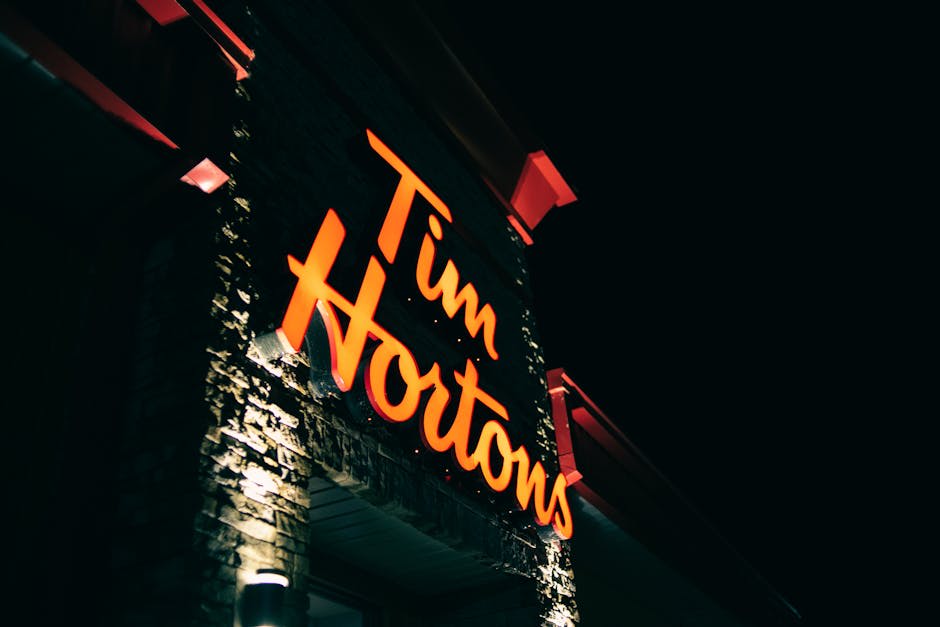 An illuminated Tim Hortons sign on a brick wall photographed at night. The orange letters are brightly lit against a dark sky backdrop, highlighting the iconic branding of the coffee shop chain.