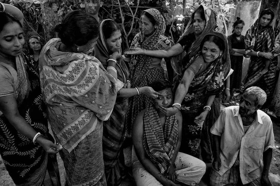 A black and white image showing a group of people participating in a traditional ceremony. Several women in saris are applying substances to a seated person's face, while others look on or assist. The scene takes place outdoors, with natural elements in the background. The participants are engaged and focused on the ceremonial activity.