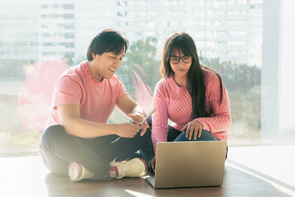A man and a woman sitting on the floor by a window, both wearing casual pink sweaters, discussing in front of a laptop. The man is holding a smartphone, and they appear to be reviewing something on the laptop's screen.