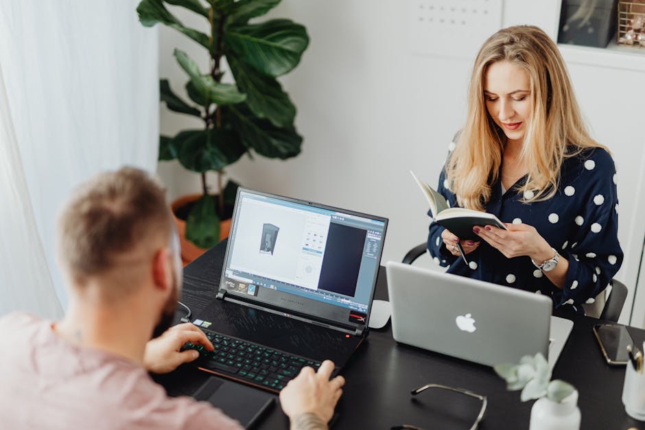 A man and a woman are working at a desk. The man is using a laptop computer while the woman is reading a notebook with her laptop open beside her. They appear to be focused and engaged with their tasks, suggesting a collaborative or office environment.