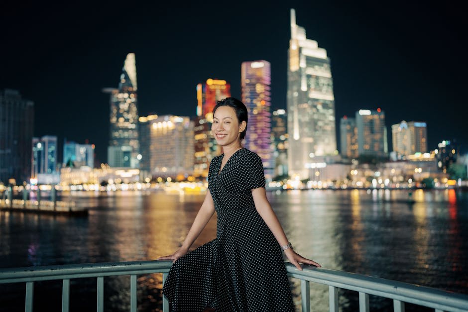 A woman in a black polka dot dress is smiling while standing on a waterfront railing at night, with illuminated city skyscrapers in the background.