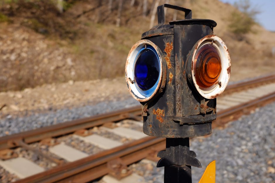 Close-up of a rusted railway signal with blue and orange lenses positioned beside train tracks in an outdoor setting.