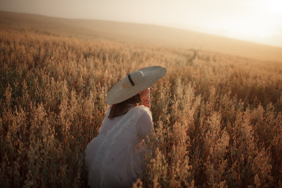 A person wearing a straw hat and white dress kneels in a sunlit field of tall grasses at sunset.
