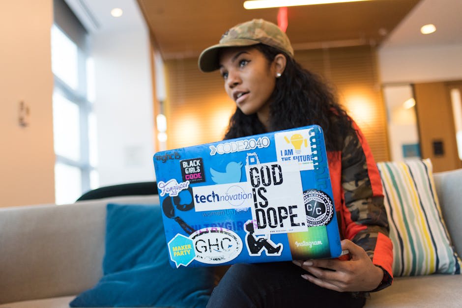 A woman wearing a camouflage jacket and cap is sitting on a couch with a colorful laptop covered in various inspirational and tech-themed stickers. The setting appears to be a modern and bright indoor space with cushions and lighting in the background. The subject seems engaged in a conversation or discussion.
