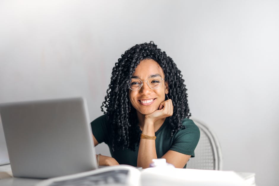 A woman with curly hair and glasses is sitting at a desk, smiling at the camera. She is in a workspace with a laptop in front of her, suggesting a work or study environment.