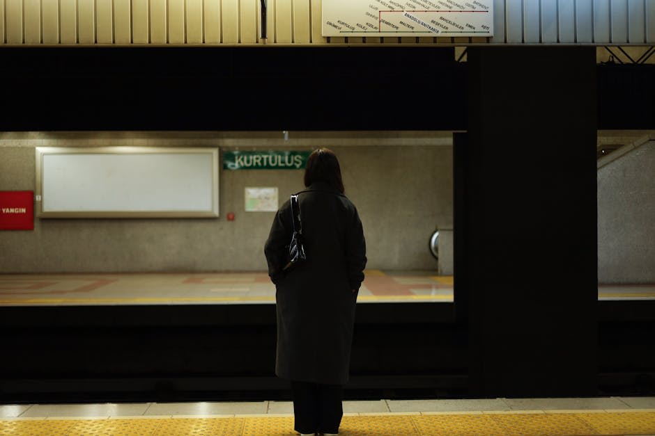 A woman standing alone on a subway platform, facing the tracks. She is wearing a dark coat and holding a bag. The setting is dimly lit, with an empty station named 'Kurtulus' in the background.